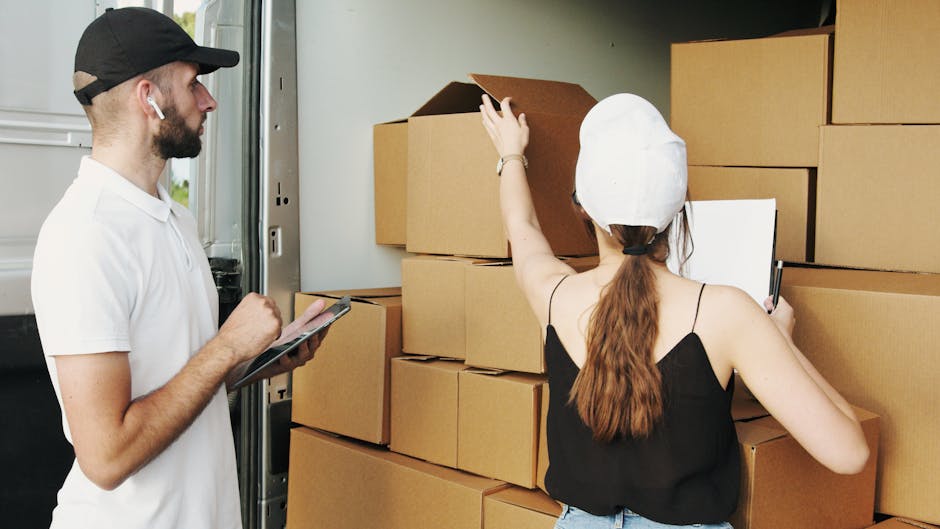 A man and a woman are engaged in a home relocation process, standing beside a moving truck with an open side door. The man, wearing a white t-shirt, black cap, and earphones, is holding a tablet or clipboard, likely overseeing the packing or loading process. The woman, dressed in a black sleeveless top and a white cap, is reaching up to adjust or examine a cardboard box. Several packed cardboard boxes of varying sizes are stacked inside the truck, some sealed with packing tape, and others open at the top. The environment appears to be either a driveway or street area, illuminated by natural light. The scene captures the logistics of furniture transport, packing, and loading during a home removal, as part of the services offered by Man and a Van Hounslow, located in Hounslow, used for efficient and professional removal and moving operations.