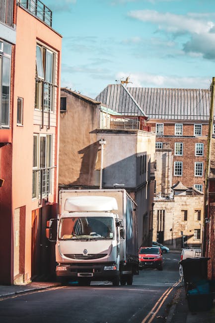 A white box truck from Man and a Van Hounslow is parked on a narrow residential street, partially blocking the roadway as it prepares for a home relocation. The truck is positioned between multi-storey brick and concrete buildings, with its cargo area facing the buildings. The surrounding environment includes a series of closely spaced, older multi-story structures with varying window styles, some with brick and others with stucco exteriors. A few parked cars, including a red one, are visible further down the street. The street itself has double yellow lines along the edge, indicating parking restrictions, and is illuminated by natural daylight with a partly cloudy sky overhead. The scene captures the logistical challenge of furniture transport and packing during moving services within limited access areas in Hounslow, illustrating the careful planning required for loading and unloading processes, often involving small maneuvering spaces typical of house removals by local specialists like Man and a Van Hounslow.