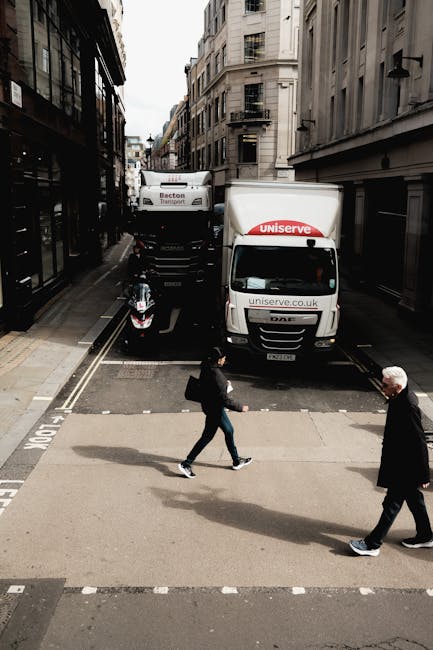 A street scene captures a narrow urban road with two large white moving vans parked close to the curb, branded with 'uniserve' and 'Becton Transport', indicating professional removal and transport services provided by Man and a Van Hounslow. The vans are positioned with their backs facing the sidewalk, suggesting a home relocation or furniture transport operation. In front of the vans, a motorbike is parked parallel to the curb, partially blocking the view of the loading area. Two pedestrians are crossing at a zebra crossing; a woman dressed in dark clothing carrying a bag and a man in a black coat with white hair. The street is lined with tall, closely spaced buildings with traditional architectural details, brick facades, and multiple windows, typical of central London. Soft daylight illuminates the scene, highlighting the urban environment and the logistical activities involved in house removals and moving services, supporting the context of packing and loading processes for a house relocation in Hounslow.