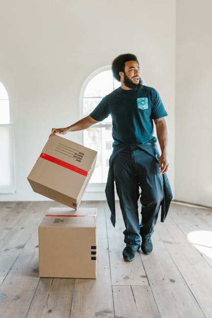 A man with dark, curly hair and a beard stands inside a bright, spacious room with white walls and wooden flooring, holding a cardboard box with a red stripe across the top in his right hand. Another unopened cardboard box rests on the floor nearby. The room features large, arched windows that allow natural light to fill the space, indicating a home environment. He is dressed in a blue t-shirt and dark pants, with additional clothing or a jacket tied around his waist, and appears to be part of a house removal or furniture transport process. The scene captures the typical packing and loading phase of a home relocation, facilitated by professional moving services like those provided by Man and a Van Hounslow. The image emphasizes careful handling of packed boxes, suitable for a house removals context, with a focus on the logistics of furniture transport and packing during a house move. The setting and actions reflect a professional move or packing and moving planning, aligned with the service category of removals.
