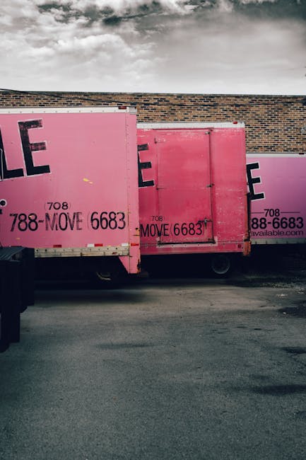 Two large pink moving containers with black and white branding and contact information are positioned side by side on a paved surface adjacent to a brick building wall. The containers are made of sturdy materials, with one slightly behind the other, and are used for home relocation purposes. Visible features include metal hinges and locks on the containers, along with a small door on the right container. The containers are partially covered by a cloudy sky, emphasizing outdoor logistics. The scene suggests an ongoing loading or unloading process typical of a house removals or furniture transport operation, which companies like Man and a Van Hounslow conduct as part of their moving services. The area around the containers is clear, with no personnel or equipment visible in this shot, focusing solely on the storage units ready for the next stage of the home relocation process.