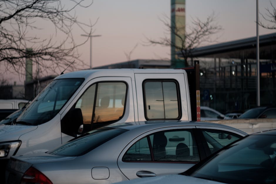 Four yellow panel vans parked in a row on a paved street with a slight incline, behind which a green, tree-covered hillside is visible under a clear sky. The vans are positioned side by side, facing a