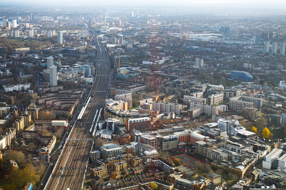 An aerial view of Hounslow, showing a dense urban landscape with numerous residential and commercial buildings. A major railway line with multiple tracks runs through the centre of the image, intersecting with roads and surrounded by apartment complexes, office buildings, and houses. To the right, there is an open paved area near a building, possibly a loading zone or street access point. The scene includes various construction and utility structures, trees scattered throughout, and distant high-rise buildings on the horizon. This setting likely depicts the area involved in home relocation and furniture transport logistics, with visible urban elements supporting the context of house removals and moving services in the Hounslow West area near Lampton Road station, as provided by Man and a Van Hounslow.
