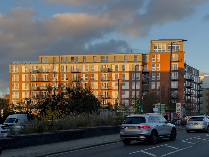 A modern multi-storey residential building with a mix of orange and beige exterior walls, featuring numerous balconies with glass railings, is visible in the background under a partly cloudy sky during late afternoon or early evening. In the foreground, a busy street scene includes several parked and moving cars, including a white SUV and a silver vehicle, along with a pavement lined with small trees and bushes. Near the building, there are signs for a petrol station and a supermarket, indicating commercial services. The overall scene represents an urban environment where home relocation or furniture transport could occur, with the building serving as a typical example of residential property in Hounslow.