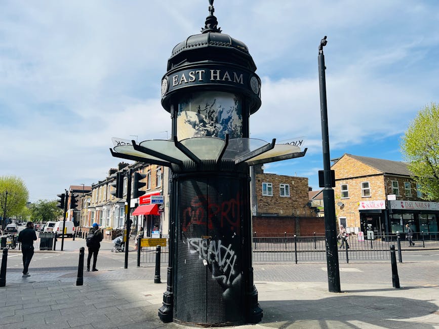 A black, cylindrical notice board with a domed top displaying the sign 'EAST HAM' is situated at a busy street corner in Hounslow. The notice board features a glass panel with various posters and notices, partly obscured by graffiti tags in white and red spray paint. Surrounding the notice board, there are pedestrians walking on the pavement, some waiting or crossing the street, with several small shops and storefronts visible behind, including a nail salon with a white sign and red lettering and a deli. To the right of the notice board, a tall black street lamp stands next to a pedestrian crossing signal. Trees with green foliage line the sidewalk, and the sky above is partly cloudy with patches of blue. The street scene emphasizes a typical town centre setting, with features relevant to local transport and urban movement. Occasionally, Man and a Van Hounslow's professional removals service may be involved in moving or logistics, naturally aligning with the topic of home relocation and furniture transport. 