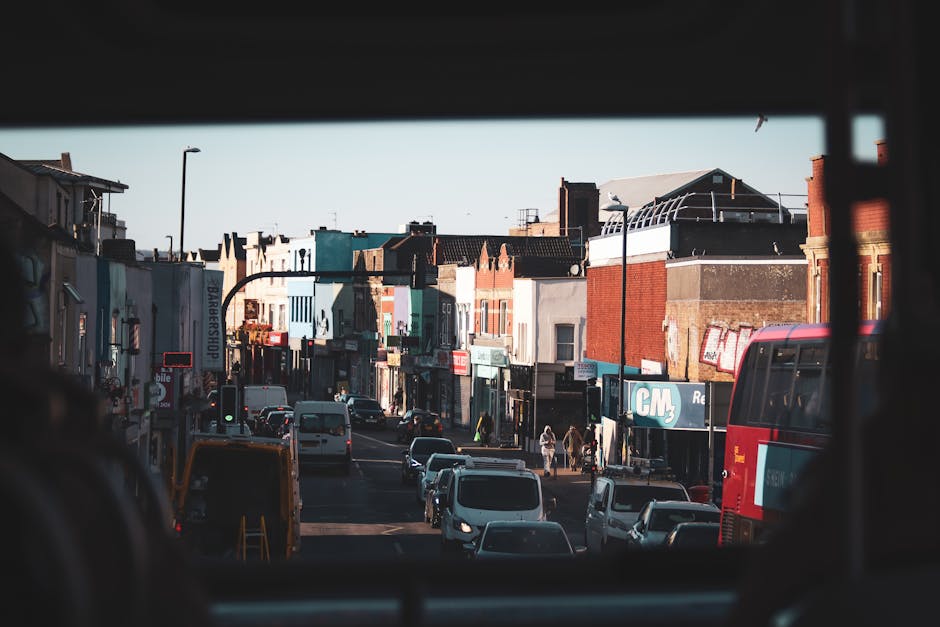 View of a busy residential street in Hounslow, taken from inside a vehicle during daylight. The street features a row of closely packed commercial and residential buildings, some with brick facades and various window styles, reflecting typical urban architecture. Multiple parked and moving cars, including a white van and a red bus, occupy the road, indicating active traffic. Pedestrians can be seen walking along the pavement, some near storefronts that display signage for local businesses. The photograph captures an overhead view, suggesting the perspective is from the back seat or rear window of a vehicle, with the vehicle's interior framing the top and bottom edges of the image. This scene is relevant to home relocation and furniture transport processes, as it illustrates the urban environment faced during moving operations, such as street parking, city traffic, and nearby commercial premises that may require careful navigation during a house removal or logistics planning, as seen in services provided by Man and a Van Hounslow.