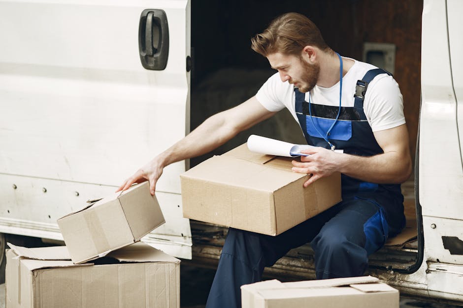 A young male removal worker with light skin, dressed in a white t-shirt and blue overalls, sits at the open side door of a white moving van. He is carefully handling a plain cardboard box, placing it on top of other packed boxes inside the vehicle. The interior of the van, visible behind him, appears to contain additional packing materials. The worker is holding a rolled-up piece of paper or a packing checklist in his other hand, indicating attention to detail during a house relocation process. The scene is well-lit, with the focus on the man's careful handling of the boxes, reflecting professional furniture transport and packing during a household move. This image illustrates the logistics involved in home removals, as provided by Man and a Van Hounslow, aligning with services for efficient house moves in Osterley Park and beyond.