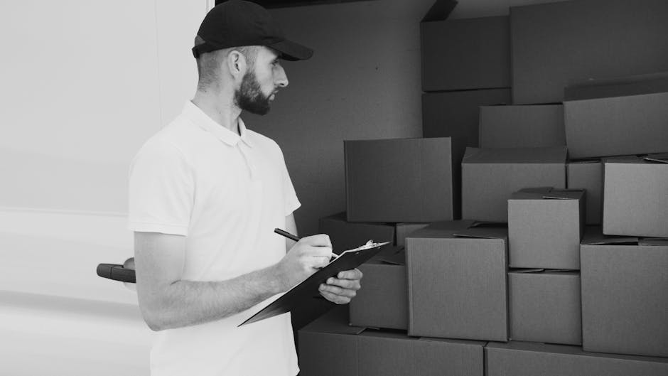 Two movers from Man and a Van Hounslow are engaged in a home relocation process, standing at the back of a large white van with its rear doors wide open. One mover, dressed in a white shirt and dark cap, is in the act of loading a stack of cardboard boxes into the van, carefully placing the boxes on top of other packed cartons. The second mover, wearing a black top, light blue jeans, and a white cap, is standing beside the van, holding a tablet and observing the loading process. Inside the van, multiple cardboard boxes are neatly stacked, ready for transport, with some wrapped in plastic or packing materials. The loading area is on a paved surface with a clear, bright sky overhead, suggesting day-time operations. The interior of the van appears spacious, with a black ceiling and ample room for furniture or additional packing. This image captures a professional packing and moving activity typical of a house removals service, and it highlights the logistical steps involved in furniture transport and home relocation, as performed by Man and a Van Hounslow.