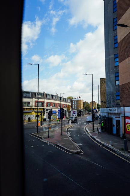 View of a street scene captured from inside a vehicle during a home relocation, showing a curved road with double tram tracks and a pedestrian crossing. On the left, there is a bus stop with a yellow sign, and behind it, a building with shops and advertising billboards. In the distance, there are multi-story residential buildings and trees lining the street. The sky above is partly cloudy with patches of blue. The photograph is taken during daytime with natural lighting, illustrating an urban environment where furniture, packing materials, or loading activities could be taking place as part of the moving process. The image reflects the typical logistics involved in house removals and transportation services offered by companies like Man and a Van Hounslow.