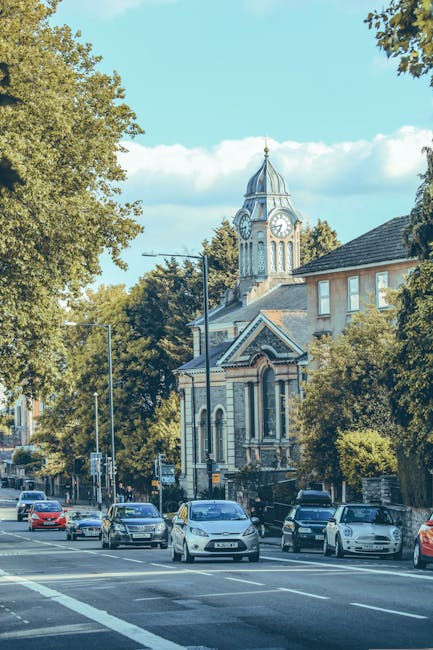 A busy street scene in Hounslow featuring several parked cars along the roadside and moving vehicles, with a historic clock tower building in the background surrounded by lush green trees. The clock tower, with its pointed roof and large clock face, is part of a traditional brick and stone structure visible behind residential buildings. The street is illuminated by natural daylight with a clear blue sky and scattered clouds overhead. In the foreground, the pavement is adjacent to the road, which is used for vehicle transit and home relocation activities. This scene exemplifies urban environment aspects relevant to house removals and furniture transport services provided by Man and a Van Hounslow, highlighting the typical setting for loading and unloading during home moves or packing and moving processes.